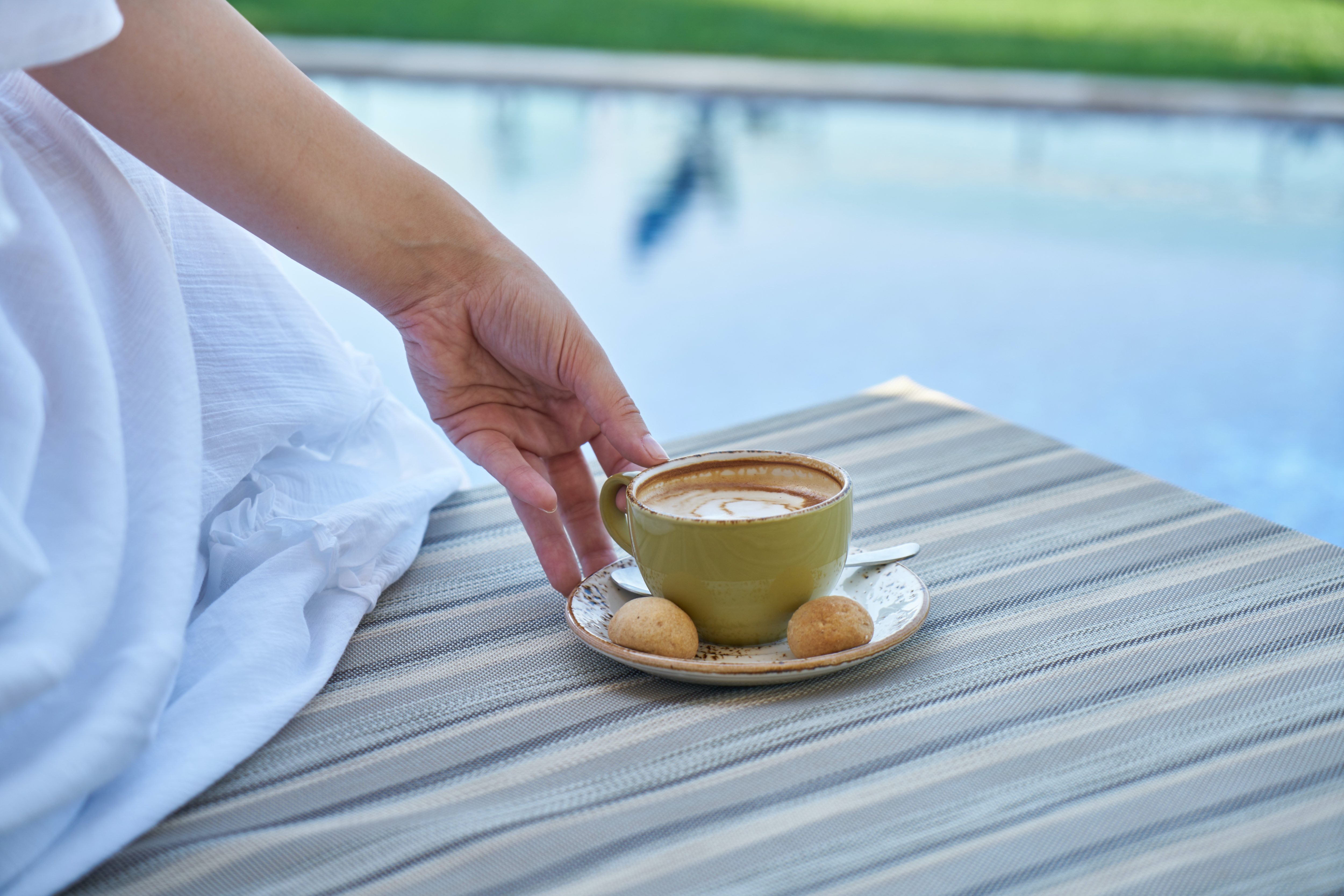 pexels-engin-akyurt-2861351 En hand sträcks fram mot en kopp kaffe som står på kanten av en solstol vid en pool.
